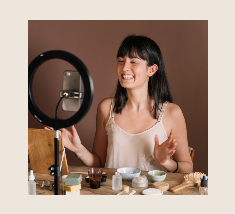 By Ivelis Social Agency, Woman creating social media content with a ring light, phones, and products on her desk